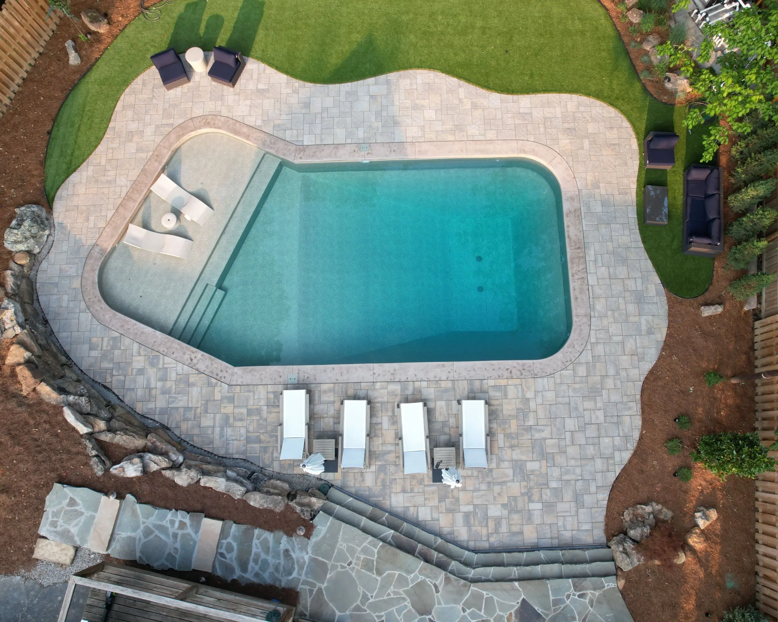 Aerial view of a Louisville backyard featuring a rectangular swimming pool surrounded by stone pavers. Six lounge chairs line one side, with lush green turf and shrubs in the background. Two dark outdoor sofas with a table are on the opposite end.