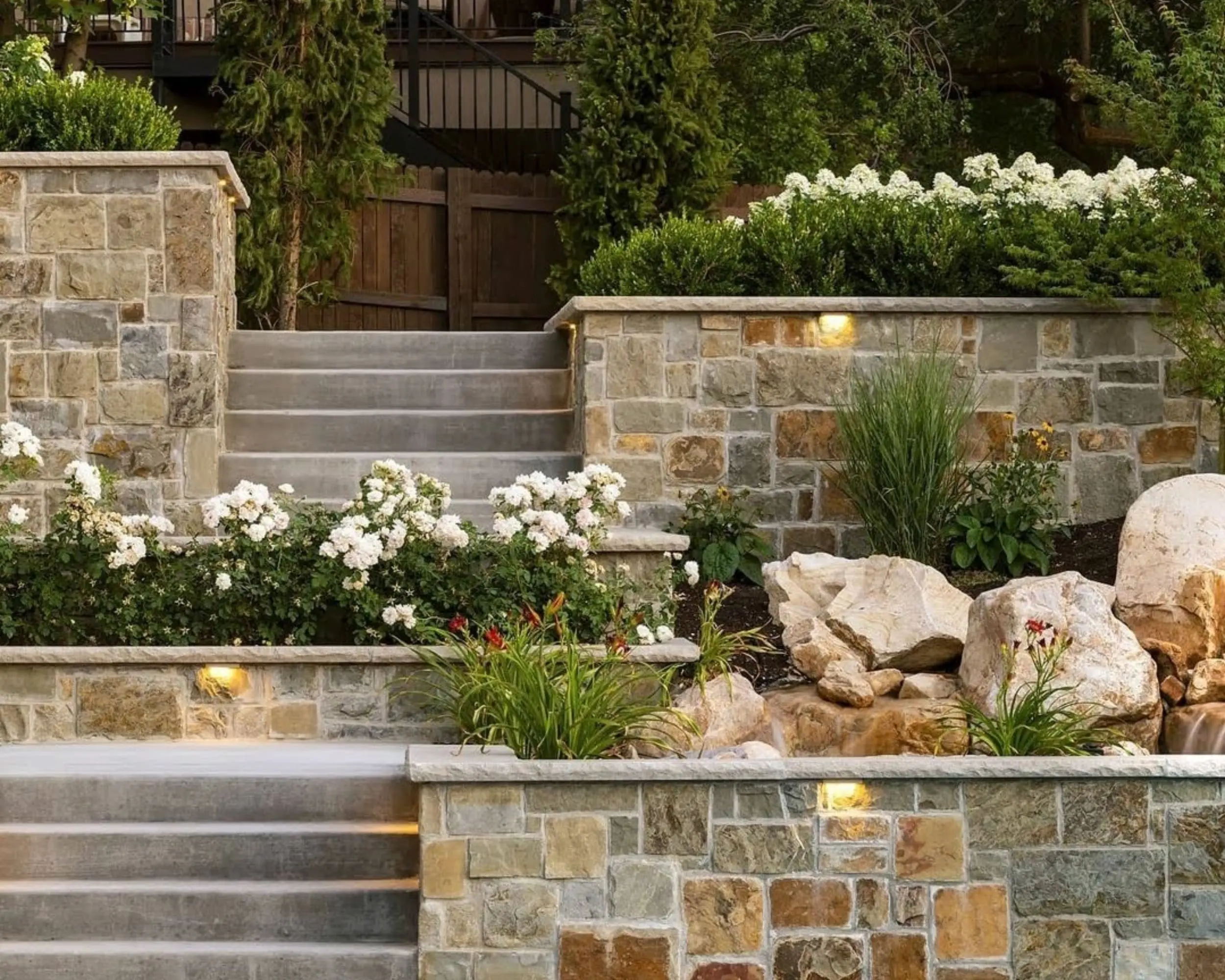 The charming stone garden staircase in this Louisville landscape features flower beds with white roses and green shrubs, flanked by stone walls. Large rocks rest near vibrant red flowers, while soft lighting gracefully illuminates the Kentucky garden path.